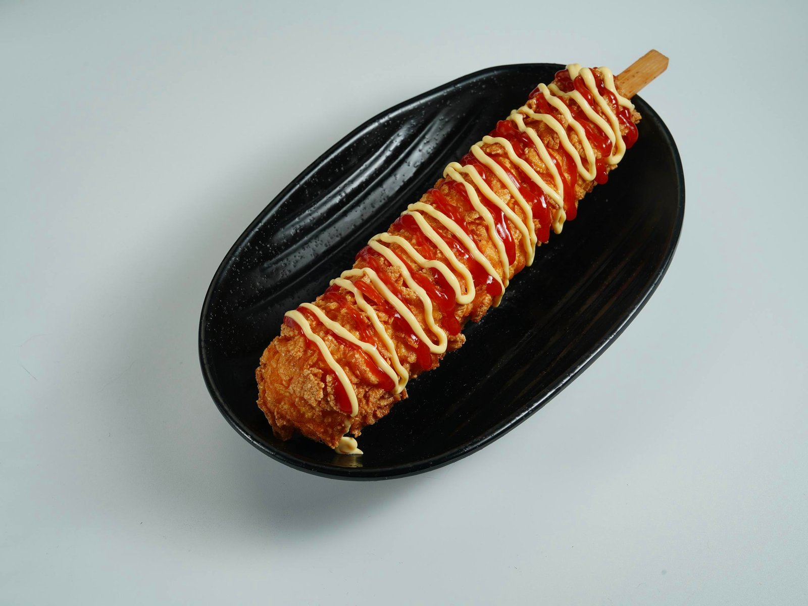 Close-up of a Korean corn dog drizzled with ketchup and mustard on a black plate.