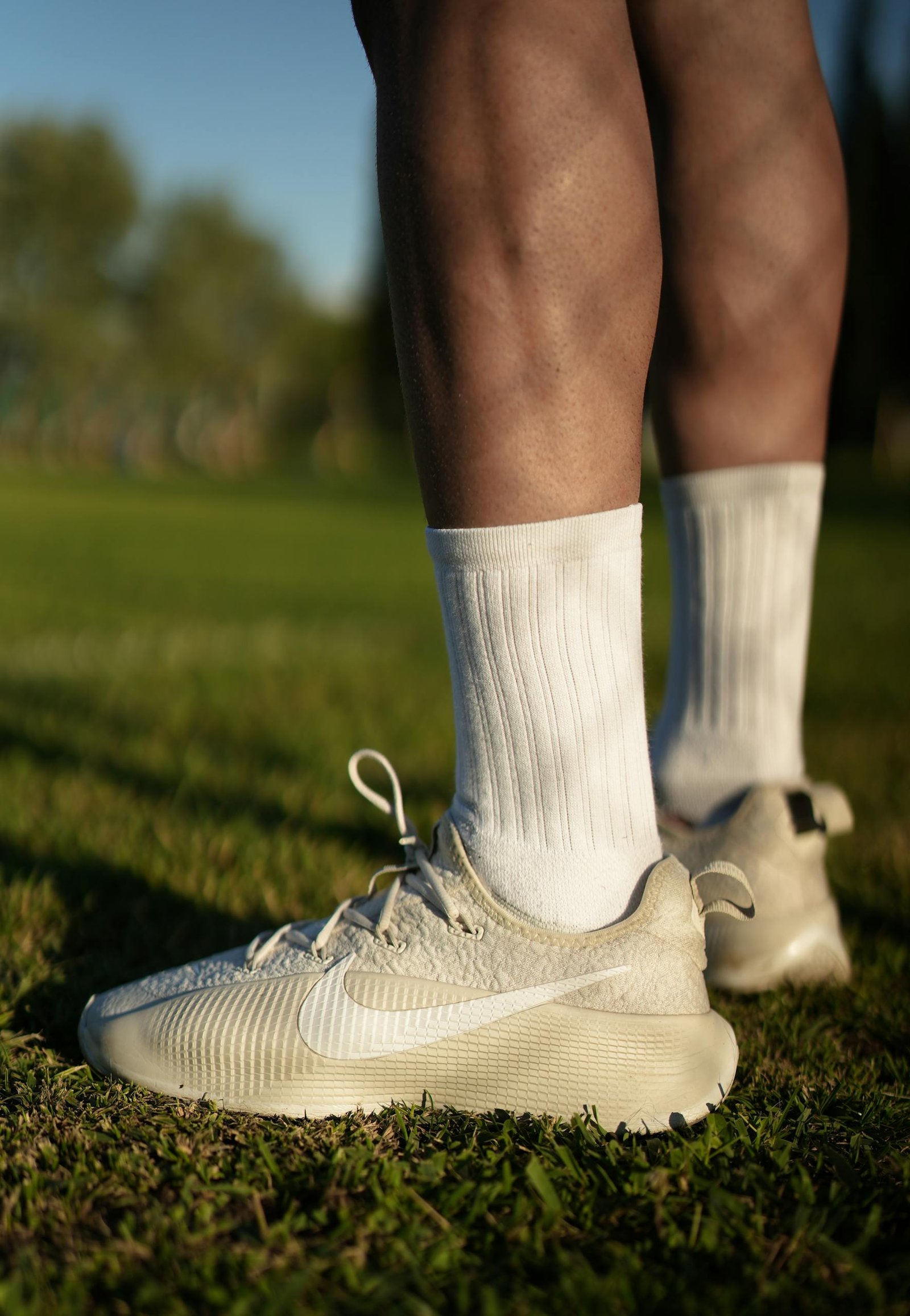 Close-up of legs in white Nike sneakers and socks standing on grass, sunny day.